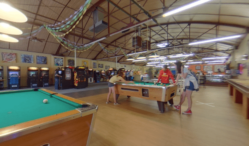 A lively arcade with pool tables and vintage games, featuring people enjoying their time in a spacious, colorful setting.