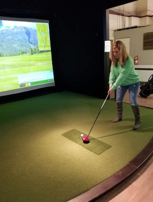 A woman in a green sweater prepares to tee off in a golf simulator, with a screen displaying a scenic course in the background.