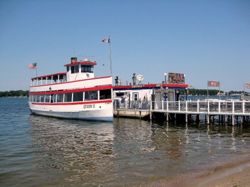 A white and red boat docked at a pier on a calm lake, with a bar and people visible on the deck.