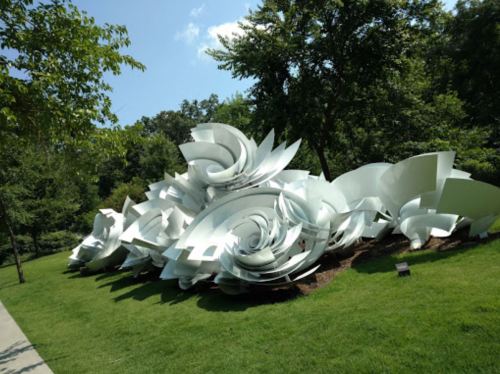 Abstract white sculpture resembling swirling petals, set in a green park with trees and blue sky in the background.