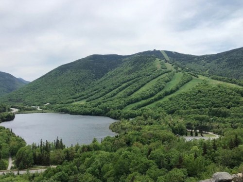 A scenic view of green mountains and a calm lake under a cloudy sky, with ski slopes visible on the hillside.
