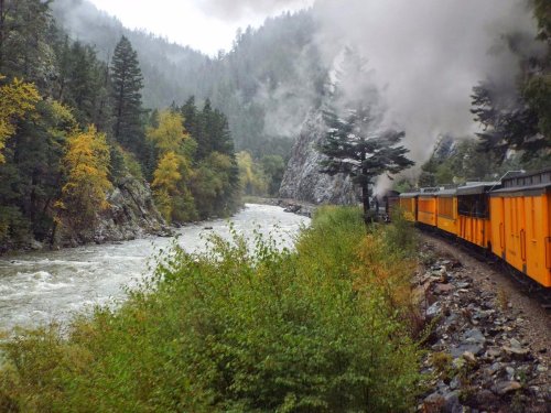 A steam train travels alongside a river, surrounded by lush greenery and misty mountains on a rainy day.