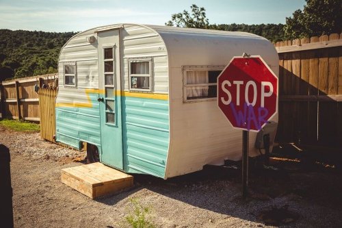 A vintage camper trailer beside a wooden fence, with a graffiti-covered stop sign reading "STOP WAR."