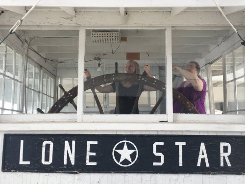 Two women steer a ship from the wheelhouse, with a sign reading "LONE STAR" visible in front of them.