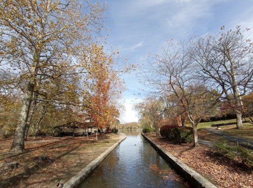 A serene park scene with a calm waterway, surrounded by trees with autumn leaves under a clear blue sky.