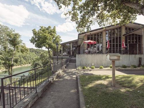 A modern building with large windows overlooks a river, surrounded by trees and outdoor seating with red umbrellas.
