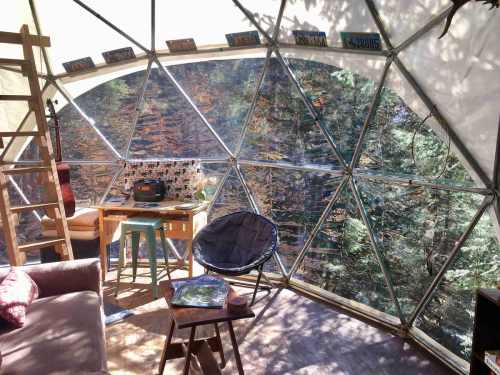 Interior of a geodesic dome with a wooden table, chairs, and large windows showcasing a forest view.