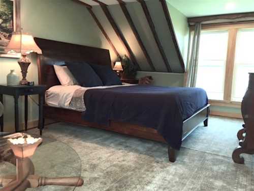 Cozy bedroom with a wooden bed, blue bedding, and sloped ceiling, featuring a glass table and soft lighting.