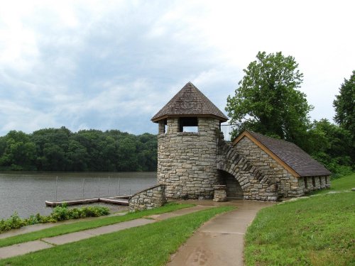 A stone tower with a sloped roof beside a calm lake, surrounded by green trees and a paved walkway.