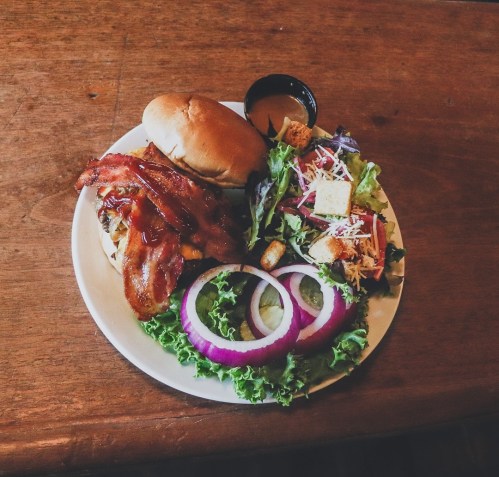 A plate with a bacon burger, fresh salad with croutons, and a side of dressing on a wooden table.