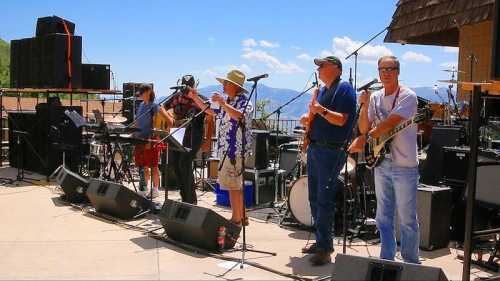 A band performs on an outdoor stage with mountains in the background, surrounded by musical equipment and instruments.