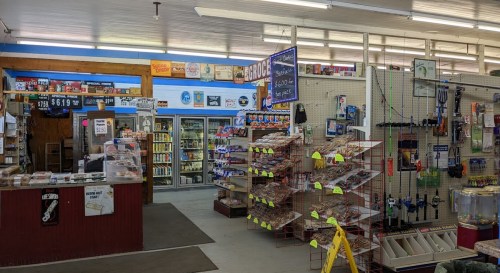 Interior of a convenience store featuring shelves of snacks, drinks, and various tools, with a checkout counter visible.