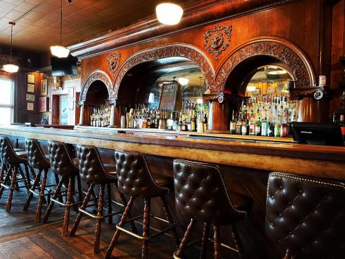 A wooden bar with ornate arches, lined with leather-tufted stools and shelves of various bottles in a cozy setting.