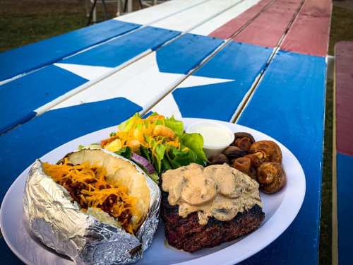A plate of food featuring a baked potato, salad, mushrooms, and a steak on a table painted with a Texas flag design.