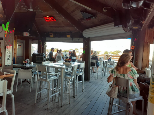 A lively bar interior with wooden decor, tables, and patrons enjoying drinks and conversation by large windows.