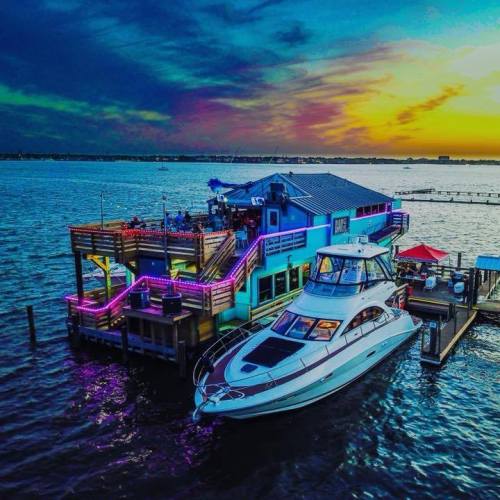 A vibrant waterfront restaurant with colorful lights, a docked yacht, and a stunning sunset over the water.