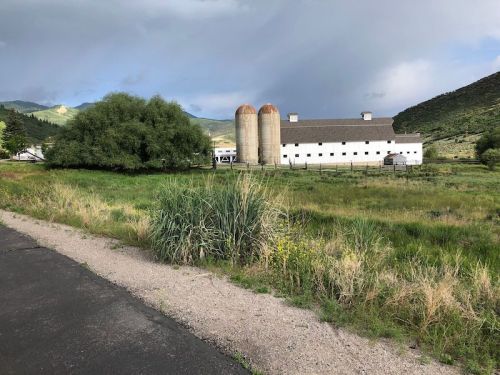 A white barn with silos surrounded by green fields and mountains under a cloudy sky.