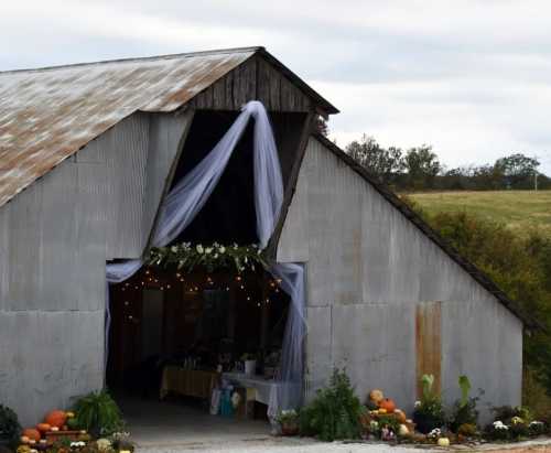 A rustic barn entrance adorned with draped fabric, flowers, and pumpkins, set against a cloudy sky.