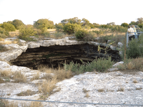 A large cave entrance surrounded by dry grass and shrubs, with people standing nearby on a viewing platform.