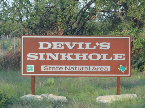Sign for Devil's Sinkhole State Natural Area in Texas, featuring brown background and white text. Green state park logo included.