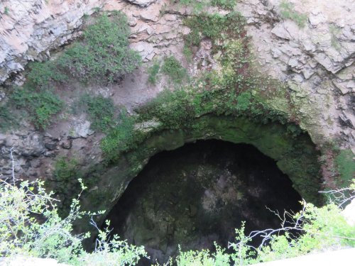 A deep, circular sinkhole surrounded by rocky terrain and sparse greenery.