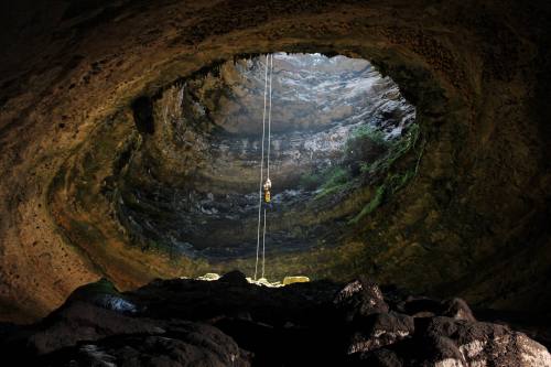 A person rappelling down into a large cave with a circular opening, illuminated by natural light from above.