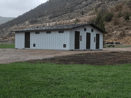 A small, gray building with multiple doors, set in a grassy area near a hillside.