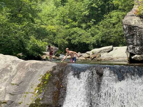 Two people wade in a shallow stream near a waterfall, surrounded by lush greenery and rocky terrain.