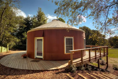 A round, brown yurt with a wooden deck, surrounded by trees and a clear blue sky.