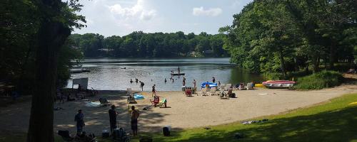 A sandy beach by a lake with people swimming, lounging, and enjoying a sunny day surrounded by trees.