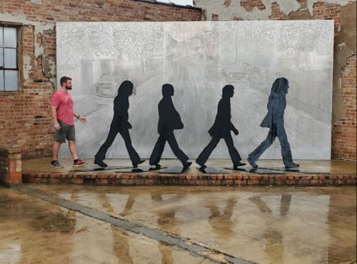A man walks past a mural depicting silhouettes of people walking in a line against a textured brick wall.