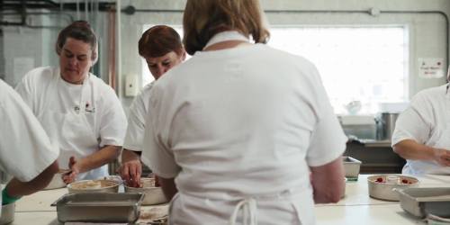 A group of women in white shirts working together in a kitchen, preparing food at a large table.