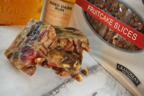 A close-up of fruitcake slices with nuts, next to a bottle of rum and a box labeled "Grandma's Fruitcake Slices."