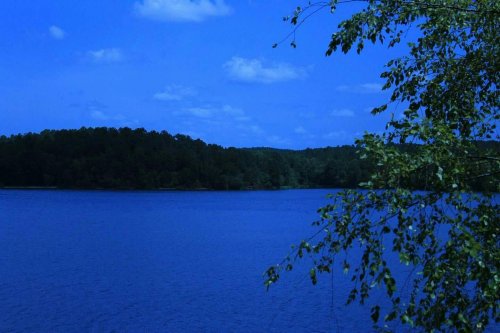 A serene blue lake surrounded by trees under a clear sky with scattered clouds.