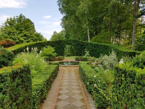 A serene garden path lined with lush greenery, leading to a tranquil pond under a leafy archway.