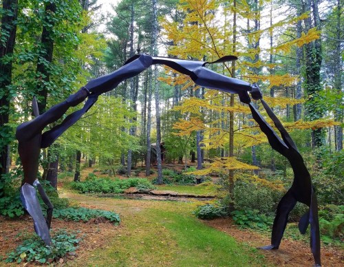 A metal sculpture of two abstract figures reaching towards each other, set in a vibrant forest with autumn foliage.