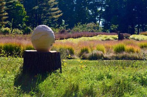 A large, white spherical sculpture sits on a tree stump in a grassy field with colorful plants in the background.