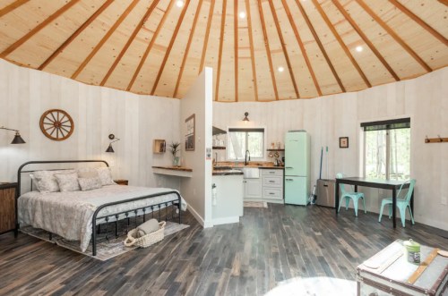 Cozy circular cabin interior with a bed, kitchen area, and vintage decor, featuring wooden beams and large windows.