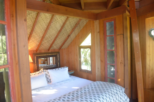 Cozy wooden bedroom with a sloped ceiling, large windows, and a neatly made bed with decorative pillows.