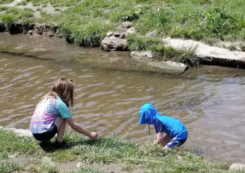 Two children play by a shallow stream, one reaching into the water while the other sits on the grass nearby.