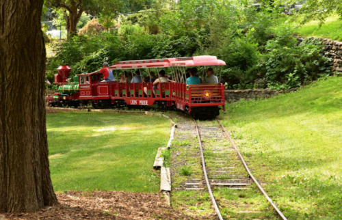 A red miniature train travels along a winding track through a lush green park. Passengers enjoy the scenic ride.