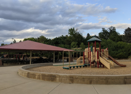 A playground with slides and climbing structures, surrounded by a gravel area and a covered picnic shelter.