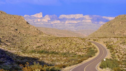 A winding road leads through arid hills, with distant mountains under a blue sky in the background.