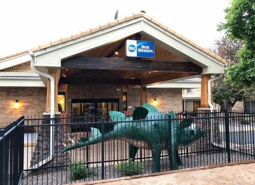 A Best Western hotel entrance featuring a large green dinosaur sculpture in front, surrounded by a fence.