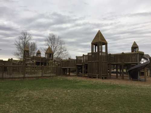 A large wooden playground with towers and a slide, set against a cloudy sky and grassy area.