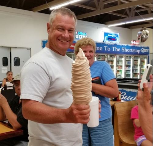 A man proudly holds a large swirl of soft serve ice cream, while a woman smiles in the background at an ice cream shop.