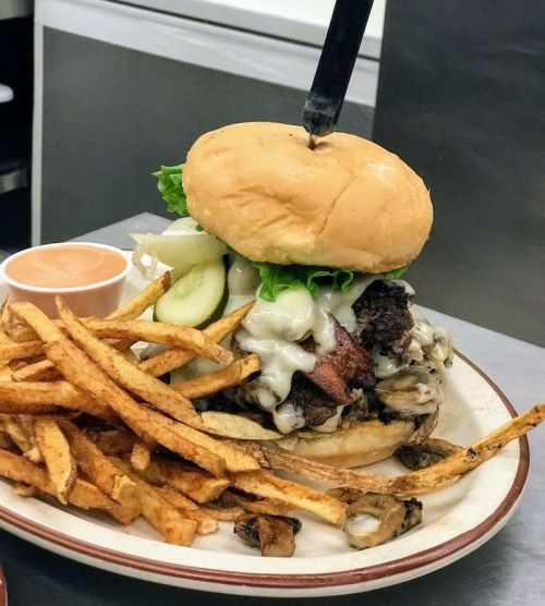 A towering burger with lettuce, pickles, and melted cheese, served with crispy fries and a side of dipping sauce.