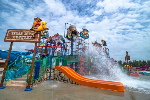 Colorful water park play area with slides, water features, and a large splash bucket under a cloudy sky.