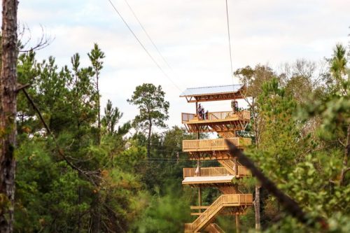 A wooden observation tower with multiple levels, surrounded by trees and a zip line in the background.