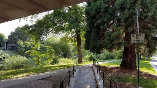 A pathway with a ramp leads to a park area, surrounded by trees and greenery, with a sign stating "No Bikes on Sidewalk."